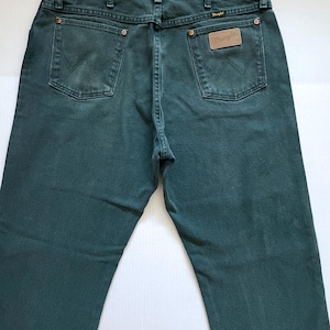 May include: Dark teal denim jeans with classic five-pocket styling. Features the Wrangler logo on a leather patch and back pockets. The jeans have a straight leg fit and are made of durable denim.