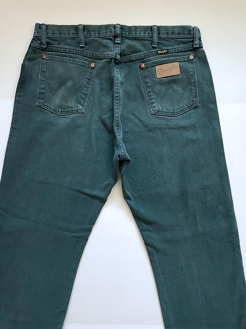 May include: Dark teal denim jeans with classic five-pocket styling. Features the Wrangler logo on a leather patch and back pockets. The jeans have a straight leg fit and are made of durable denim.