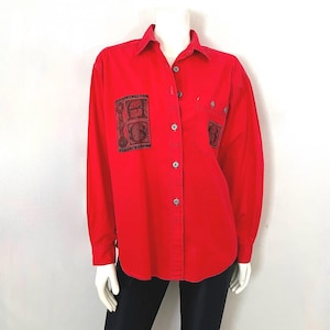 May include: A vibrant red button-down shirt with long sleeves. The shirt has two black square patches with detailed designs on the front and a single pocket. The shirt is displayed on a mannequin with black trousers.