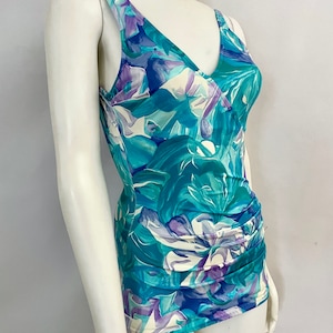 May include: A sleeveless top with a V-neckline and a vibrant floral pattern in shades of teal, blue, and purple. The fabric is ruched at the waist, creating a flattering silhouette. The top is displayed on a mannequin.