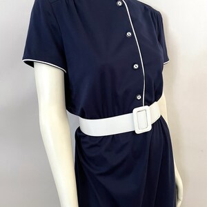 Op de afbeelding: Een marineblauwe jurk met korte mouwen, witte bies en een witte riem. De jurk heeft een knoopsluiting met witte knopen en een bijpassende witte riem met een rechthoekige gesp. De jurk wordt op een mannequin gepresenteerd.