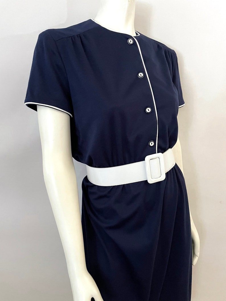 Op de afbeelding: Een marineblauwe jurk met korte mouwen, witte bies en een witte riem. De jurk heeft een knoopsluiting met witte knopen en een bijpassende witte riem met een rechthoekige gesp. De jurk wordt op een mannequin gepresenteerd.