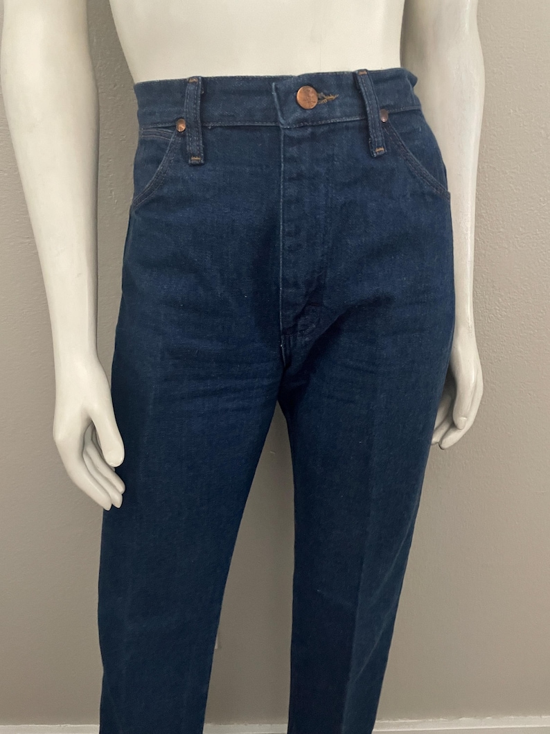 May include: Dark blue denim jeans with a classic five-pocket design. The jeans have a button and zipper closure, belt loops, and copper-colored rivets. The straight-leg style is displayed on a mannequin.