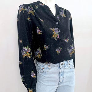 May include: Black long-sleeve blouse with a V-neck and button closure. The blouse features a floral pattern in shades of purple, yellow, and white, with small white polka dots. The blouse is paired with light blue denim jeans.