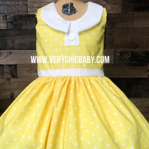 May include: A yellow dress with white polka dots and a white Peter Pan collar. The dress has a white sash and white lace trim at the hem. The dress is on a mannequin and the website www.verychicbaby.com is visible on the dress.