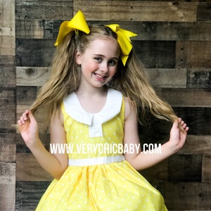 May include: A young girl wearing a yellow dress with white polka dots and a white collar. She has yellow bows in her hair and is smiling. The dress has a white lace trim and a white sash. The website www.verychicbaby.com is visible on the dress.
