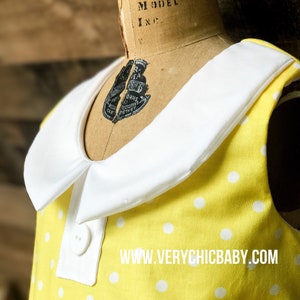 May include: A yellow sleeveless dress with white polka dots and a white Peter Pan collar. The dress has two white buttons on the front. The dress has the text "www.verychicbaby.com" printed on the front.