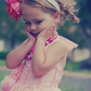May include: A young girl with curly hair wears a pink dress with a white headband and a pink beaded necklace. The girl is looking down and has her hands on her cheeks. The background is blurred and out of focus.