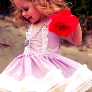 May include: A young girl wearing a lavender dress with a white lace trim and a white petticoat. She is wearing a silver tiara and a pearl necklace. She is holding a red rose in her right hand. The image has the text "Very Chic Baby Designs" in pink.