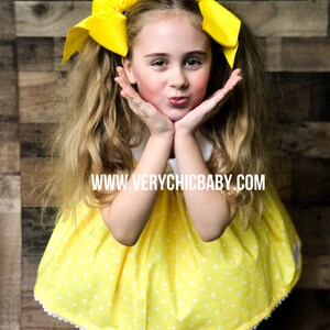 May include: A young girl wearing a yellow dress with white polka dots and a white lace trim. She is wearing two large yellow bows in her hair. The dress has a white Peter Pan collar. The girl is smiling and has her hands on her cheeks. The text "www.verychicbaby.com" is printed on the dress.