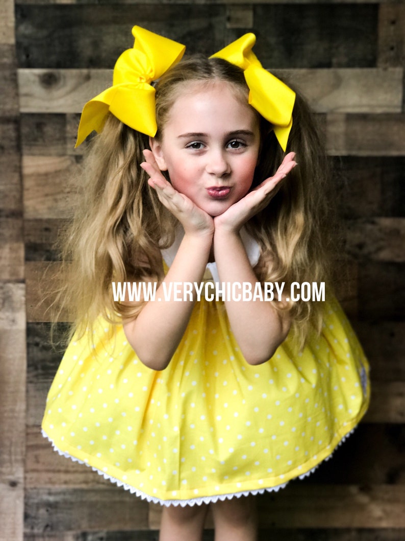 May include: A young girl wearing a yellow dress with white polka dots and a white lace trim. She is wearing two large yellow bows in her hair. The dress has a white Peter Pan collar. The girl is smiling and has her hands on her cheeks. The text "www.verychicbaby.com" is printed on the dress.