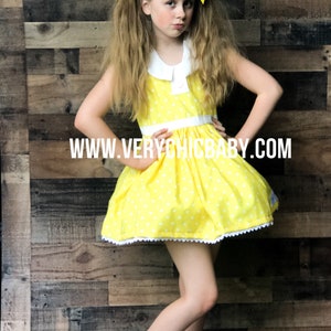 May include: A young girl wearing a yellow polka dot dress with a white collar and white trim. She has yellow bows in her hair and is wearing white shoes. The dress has a white lace trim around the bottom. The background is a wood panel wall. The text "WWW.VERYCHICBABY.COM" is printed on the dress.