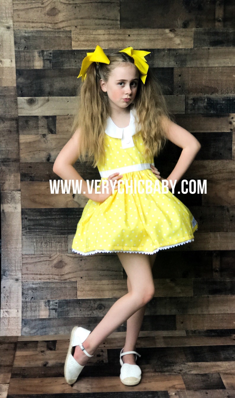 May include: A young girl wearing a yellow polka dot dress with a white collar and white trim. She has yellow bows in her hair and is wearing white shoes. The dress has a white lace trim around the bottom. The background is a wood panel wall. The text "WWW.VERYCHICBABY.COM" is printed on the dress.
