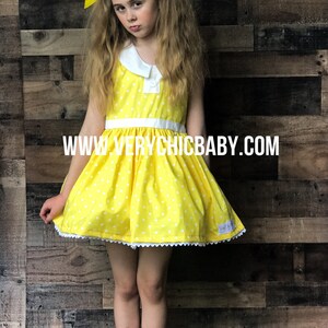 May include: A young girl wearing a yellow polka dot dress with a white collar and white trim. She is wearing white shoes and has yellow bows in her hair. The dress has a white belt and the text "www.verychicbaby.com" is printed on the front.