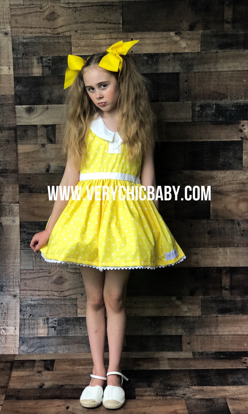 May include: A young girl wearing a yellow polka dot dress with a white collar and white trim. She is wearing white shoes and has yellow bows in her hair. The dress has a white belt and the text "www.verychicbaby.com" is printed on the front.