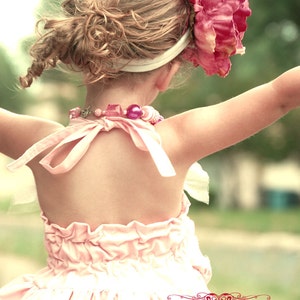 May include: A young girl wearing a pink ruffled dress with a pink flower headband and a pink beaded necklace. The girl is standing with her back to the camera and her arms outstretched. The image is in a vintage style.