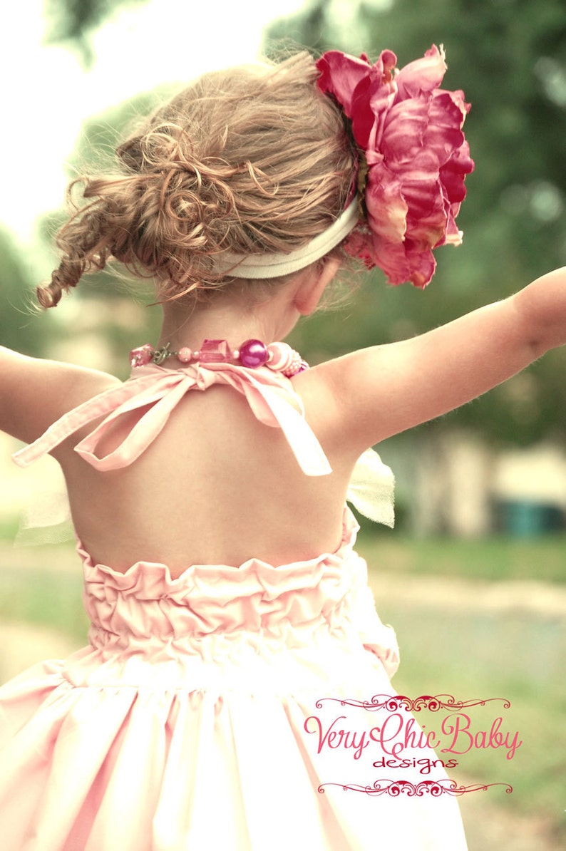 May include: A young girl wearing a pink ruffled dress with a pink flower headband and a pink beaded necklace. The girl is standing with her back to the camera and her arms outstretched. The image is in a vintage style.