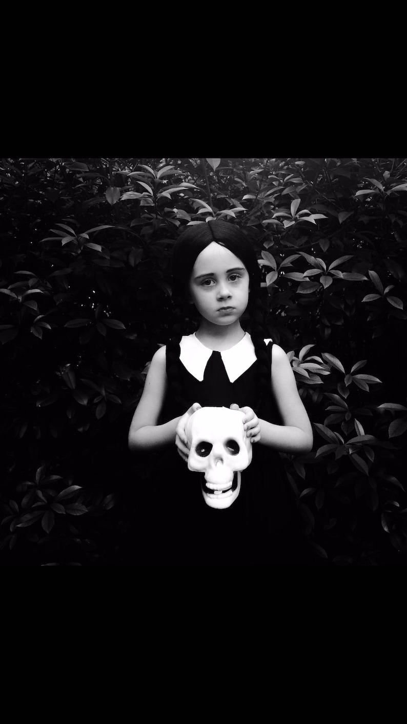 May include: A young girl in a black dress with a white collar holds a white plastic skull in front of her. She has dark hair and is standing in front of a dark green leafy background.