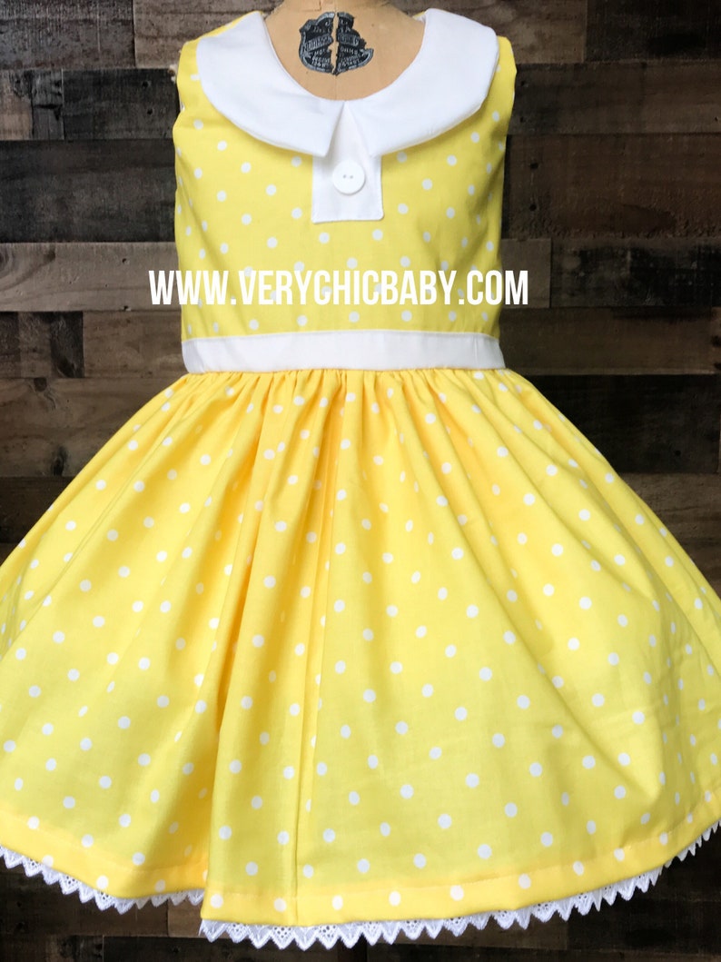 May include: A yellow dress with white polka dots and a white Peter Pan collar. The dress has a white sash and white lace trim at the hem. The dress is on a mannequin and the website www.verychicbaby.com is visible on the dress.
