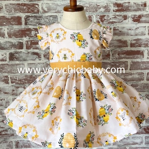 May include: A light pink dress with a repeating pattern of yellow beehives, black and yellow bees, and floral wreaths. The dress features flutter sleeves, a round neckline, and a solid mustard yellow waistband. The text www.verychicbaby.com is visible.