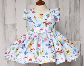 May include: A light blue dress with a Winnie the Pooh and friends print. The dress has a round neckline, short ruffle sleeves, and a full skirt. The print features Winnie the Pooh, Tigger, Piglet, and Eeyore flying kites and holding balloons.