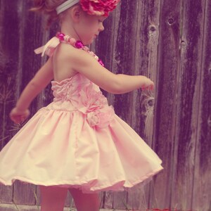 May include: A young girl wearing a pink dress with a large pink flower headband. The dress has a ruffled skirt and a flower accent on the side. The girl is standing in front of a wooden fence.
