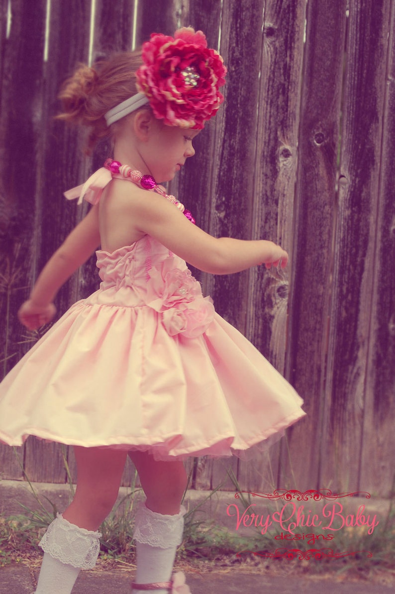 May include: A young girl wearing a pink dress with a large pink flower headband. The dress has a ruffled skirt and a flower accent on the side. The girl is standing in front of a wooden fence.