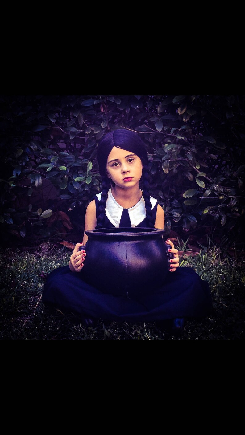 May include: A young girl dressed as a witch for Halloween, wearing a black dress with a white collar and pigtails, holds a black cauldron in front of her.