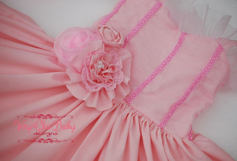 May include: A pink dress with a ruffled flower embellishment and pink lace trim. The dress has a tulle overlay and a satin bodice.