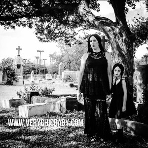 May include: Two people, a woman and a young girl, stand in a cemetery. The woman is wearing a black lace dress and the girl is wearing a black dress with a white collar. The woman has long dark hair and the girl has long dark hair with a white bow. The background is a cemetery with tombstones and trees. The text "www.verychicbaby.com" is at the bottom of the image.
