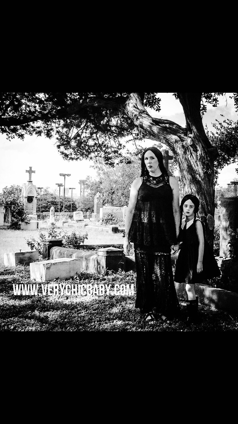 May include: Two people, a woman and a young girl, stand in a cemetery. The woman is wearing a black lace dress and the girl is wearing a black dress with a white collar. The woman has long dark hair and the girl has long dark hair with a white bow. The background is a cemetery with tombstones and trees. The text "www.verychicbaby.com" is at the bottom of the image.