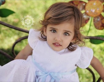 Puede incluir: Una niña pequeña con cabello castaño claro y ojos verdes que lleva un vestido blanco con ojales y una cinta azul claro. La niña está sentada en un banco de metal con un fondo verde borroso y una gran decoración floral.