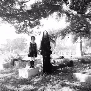 May include: Two people dressed in black clothing stand in a cemetery. The person on the left is a child wearing a black dress and white collar. The person on the right is an adult wearing a long black dress. Both are looking at the camera.