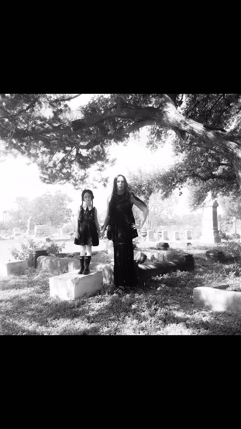 May include: Two people dressed in black clothing stand in a cemetery. The person on the left is a child wearing a black dress and white collar. The person on the right is an adult wearing a long black dress. Both are looking at the camera.