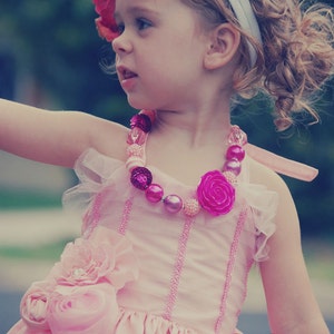 May include: A young girl wearing a pink dress with a flower embellishment and a pink and white beaded necklace. She is wearing a white headband and has curly blonde hair.