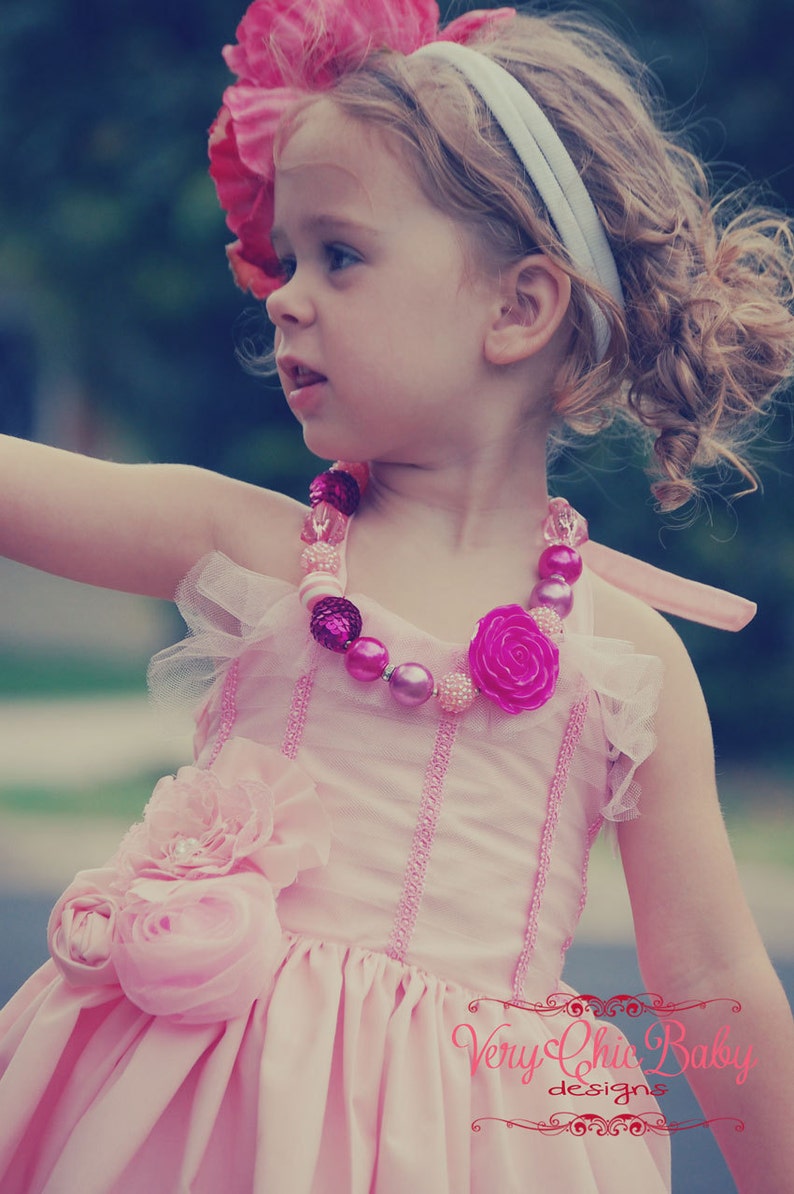 May include: A young girl wearing a pink dress with a flower embellishment and a pink and white beaded necklace. She is wearing a white headband and has curly blonde hair.