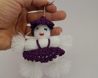 May include: A white crocheted doll keychain with purple accents. The doll has a purple hat, a purple dress, and white pom-poms for arms and legs.