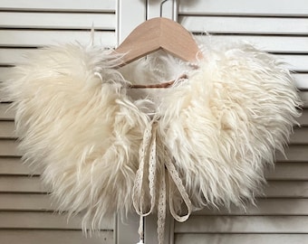 May include: A cream-colored faux fur collar featuring long, shaggy fibers and a delicate, off-white lace ribbon tie closure. The accessory is displayed on a wooden hanger, showcasing its soft, voluminous texture and light, neutral tone.