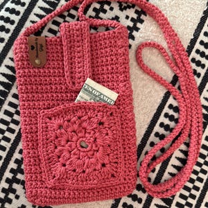 May include: A handmade, coral-colored crochet crossbody bag. The bag features a square pocket with a floral design and a long strap. A small leather tag reads "Handmade by Gigi". A dollar bill is visible in the pocket.