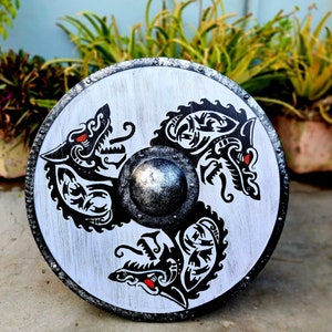 May include: A white round shield with three black wolf designs. The shield has a silver metal center and red accents in the wolf's eyes.