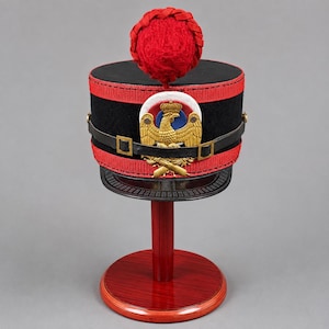 Può includere: Un berretto militare cilindrico nero con un pompon rosso brillante sulla parte superiore, fasce in tessuto rosso e un sottogola in pelle nera. Un emblema d'aquila color oro con uno sfondo circolare bianco, blu e rosso è centrato sul davanti.