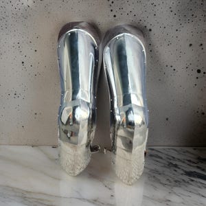 May include: A pair of polished silver-colored metal greaves designed to protect the lower legs. These armor pieces feature a smooth, reflective surface with visible rivets along the edges and a curved, anatomical shape that tapers toward the ankle.
