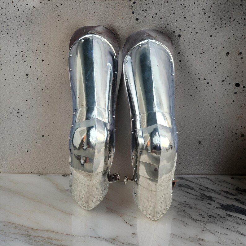 May include: A pair of polished silver-colored metal greaves designed to protect the lower legs. These armor pieces feature a smooth, reflective surface with visible rivets along the edges and a curved, anatomical shape that tapers toward the ankle.