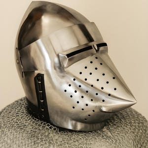 May include: A polished steel helmet with a pointed visor and numerous small ventilation holes. The helmet is part of a suit of armor and rests on a chainmail coif. The design is reminiscent of medieval knight's armor.