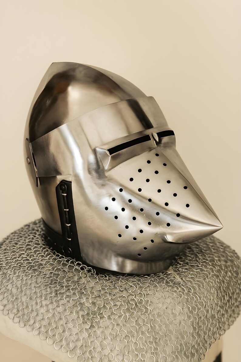 May include: A polished steel helmet with a pointed visor and numerous small ventilation holes. The helmet is part of a suit of armor and rests on a chainmail coif. The design is reminiscent of medieval knight's armor.