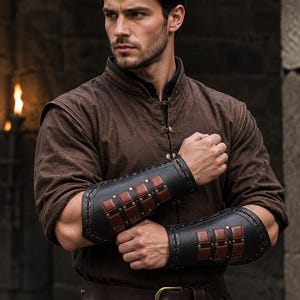 May include: A brown long-sleeved tunic with a high collar and metal fasteners, paired with black leather forearm bracers featuring square red leather accents and metal rivets, and a dark brown leather belt with a metal buckle.