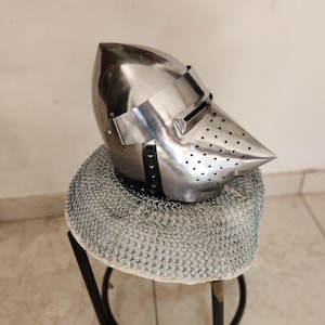 May include: A polished steel helmet with a pointed visor and small ventilation holes. The helmet rests on a chainmail coif, displayed on a small stool. The helmet's design is reminiscent of medieval armor.