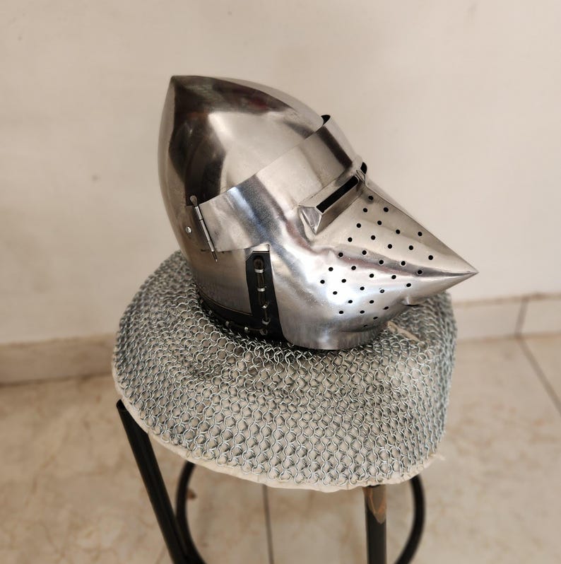 May include: A polished steel helmet with a pointed visor and small ventilation holes. The helmet rests on a chainmail coif, displayed on a small stool. The helmet's design is reminiscent of medieval armor.