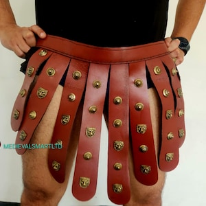 May include: A reddish-brown leather Roman skirt with brass-colored studs and decorative face emblems. The skirt is designed with vertical leather strips and a belt-like waistband, offering a historical and thematic aesthetic.