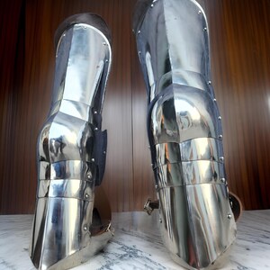 May include: A pair of polished steel greaves designed to protect the lower legs. These metallic armor pieces feature articulated plates, visible rivets, and brown leather straps with metal buckles for secure fastening around the calf area.